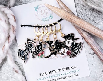 May include: Set of four black and gold metal stitch markers with different spooky Halloween designs. The designs include a skull, a crow, a witch's hat, and a plague doctor. The stitch markers are attached to gold rings. The image also includes a wooden knitting needle and a white yarn ball. The text on the card reads "The Desert Stream Craft Design Creativity www.thedesertstream.com".