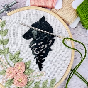 May include: A close-up of a black wolf head embroidery pattern on white fabric. The pattern is being stitched with green thread using a needle and embroidery hoop.