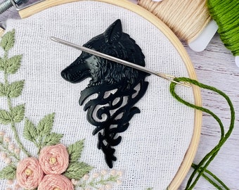 May include: A close-up of a black wolf head embroidery pattern on white fabric. The pattern is being stitched with green thread using a needle and embroidery hoop.