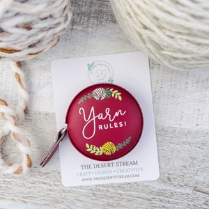 Pu&ograve; includere: Un metro a nastro rotondo bordeaux con la scritta "Yarn Rules!" in corsivo bianco. Il metro a nastro ha una piccola linguetta in pelle ed &egrave; decorato con illustrazioni di filato e foglie. Lo sfondo include gomitoli di filato e una carta bianca.