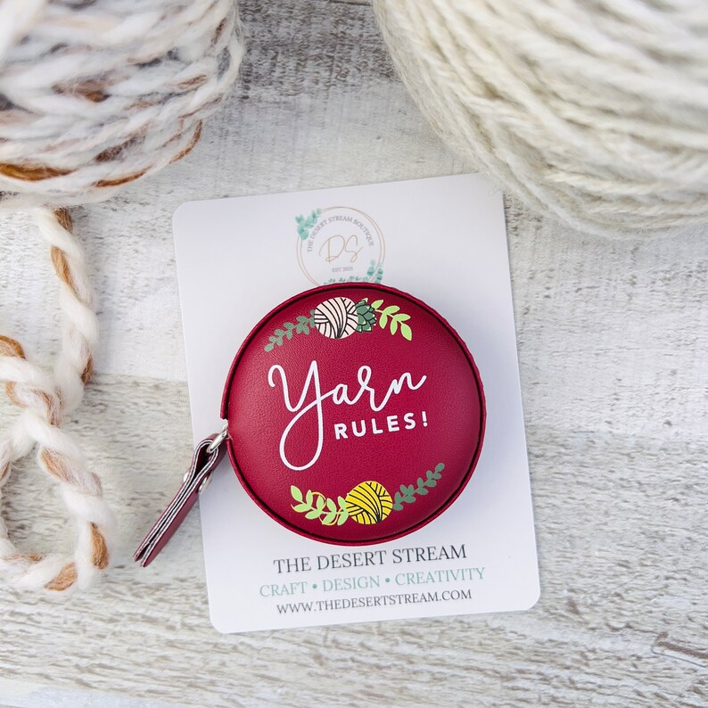 Pu&ograve; includere: Un metro a nastro rotondo bordeaux con la scritta "Yarn Rules!" in corsivo bianco. Il metro a nastro ha una piccola linguetta in pelle ed &egrave; decorato con illustrazioni di filato e foglie. Lo sfondo include gomitoli di filato e una carta bianca.