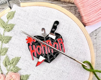 May include: A black and red heart-shaped pin with a knife and the word "Horror" in red lettering. The pin is on a white fabric background with a green embroidery design.