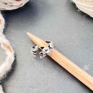 May include: A silver cat-shaped stitch marker on a wooden knitting needle. The cat is curled up and has a detailed face.