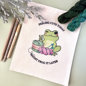 May include: A white canvas tote bag with the text "Feeling cute today, might frog it later."  A green frog with a pink and green ball of yarn is pictured.
