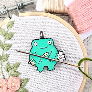 May include: A green frog holding a needle and a white flower embroidery hoop with a floral design. The frog is a needle minder, a small accessory used to hold needles while embroidering.