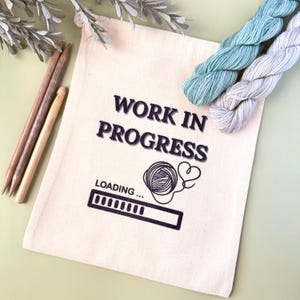May include: A white canvas bag with the text "WORK IN PROGRESS" and a loading bar graphic. The bag contains two skeins of yarn, one light blue and one white.