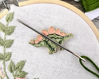 May include: A close-up of a green and pink dinosaur embroidery pattern on white fabric. A needle and thread are threaded through the fabric, ready for stitching.