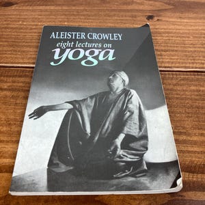 May include: A monochrome book cover with the title "ALEISTER CROWLEY eight lectures on yoga". The word "yoga" is in teal. The image shows a seated figure. The book rests on a wooden surface.