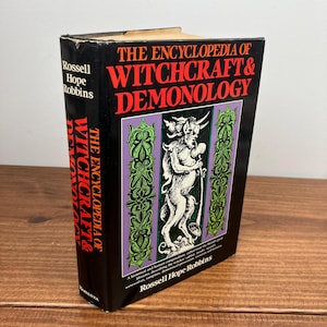Może przedstawiać: Twarda oprawa książki zatytułowana "THE ENCYCLOPEDIA OF WITCHCRAFT & DEMONOLOGY" autorstwa Rossella Hope Robbinsa. Książka ma czarną okładkę z czerwonym i białym tekstem. Okładka zawiera ilustrację demona w fioletowo-zielonej ramie.
