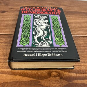Może przedstawiać: Twarda oprawa książki zatytułowanej "The Encyclopedia of Witchcraft & Demonology" z czarną okładką. Okładka zawiera centralną ilustrację rogatej postaci oraz dekoracyjne zielone i fioletowe obramowania. Widoczne jest r&oacute;wnież nazwisko autora, Rossell Hope Robbins.