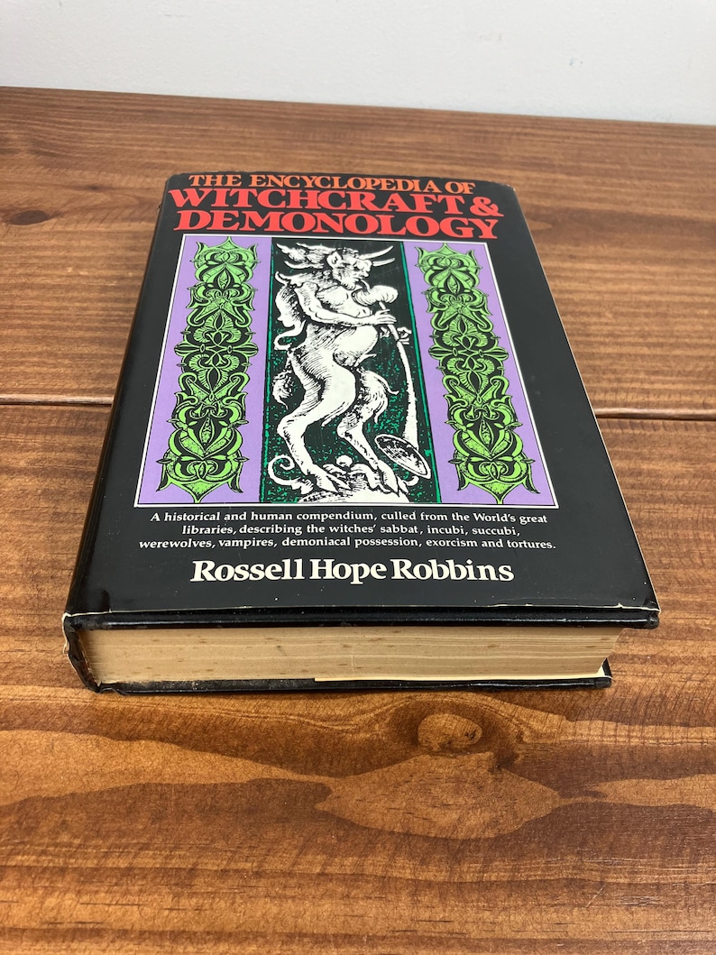 Może przedstawiać: Twarda oprawa książki zatytułowanej "The Encyclopedia of Witchcraft & Demonology" z czarną okładką. Okładka zawiera centralną ilustrację rogatej postaci oraz dekoracyjne zielone i fioletowe obramowania. Widoczne jest r&oacute;wnież nazwisko autora, Rossell Hope Robbins.