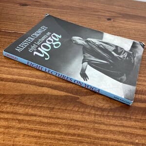 May include: A book titled "Eight Lectures on Yoga" by Aleister Crowley. The cover shows a grayscale image of a person in a yoga pose. The book has a blue spine and is on a wooden surface.