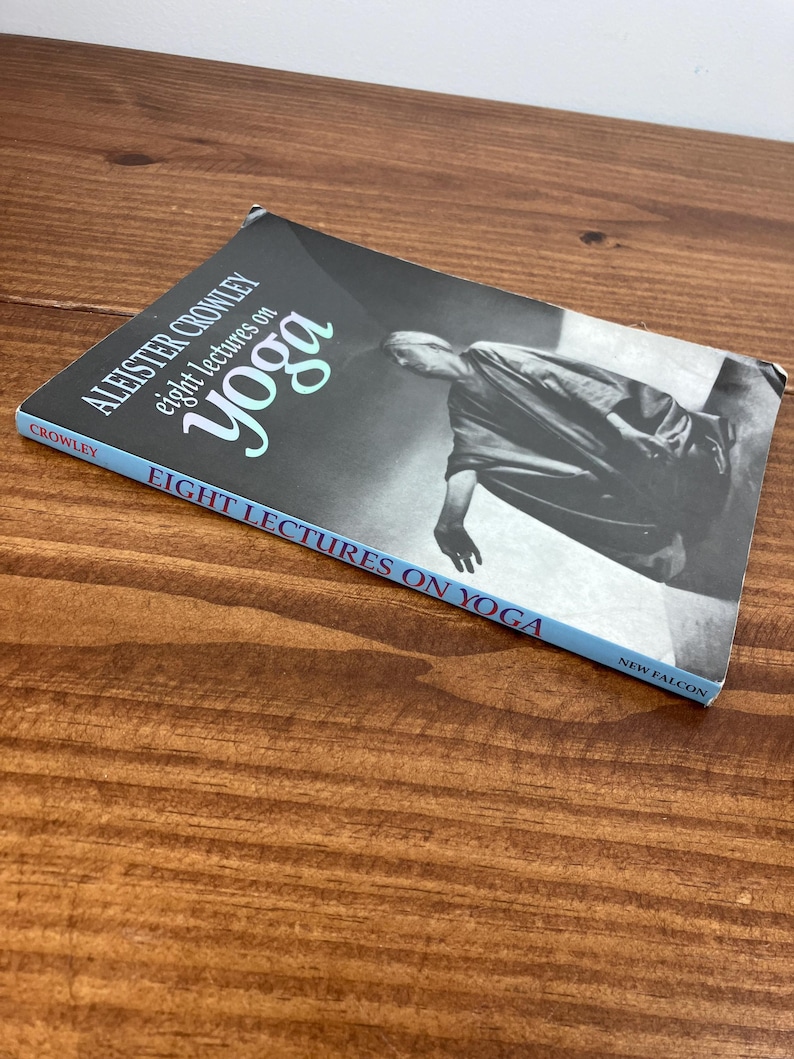 May include: A book titled "Eight Lectures on Yoga" by Aleister Crowley. The cover shows a grayscale image of a person in a yoga pose. The book has a blue spine and is on a wooden surface.