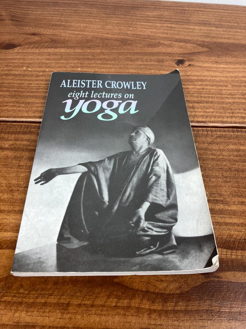 May include: A monochrome book cover with the title "ALEISTER CROWLEY eight lectures on yoga". The word "yoga" is in teal. The image shows a seated figure. The book rests on a wooden surface.