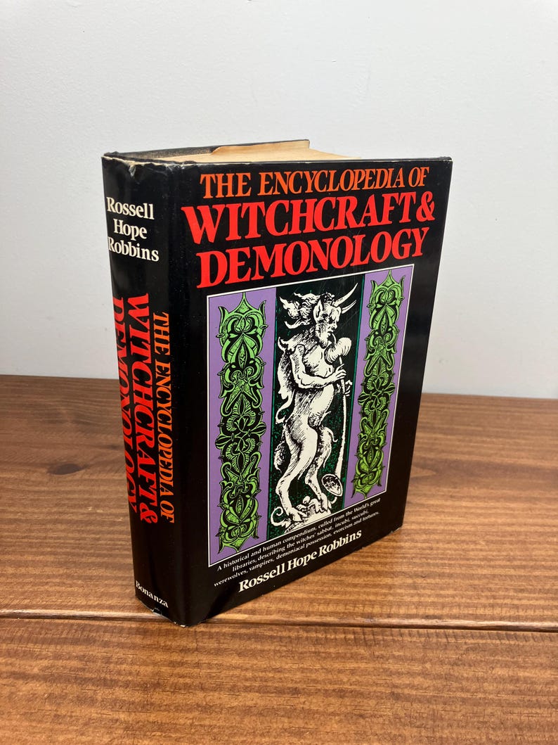 Może przedstawiać: Twarda oprawa książki zatytułowana "THE ENCYCLOPEDIA OF WITCHCRAFT & DEMONOLOGY" autorstwa Rossella Hope Robbinsa. Książka ma czarną okładkę z czerwonym i białym tekstem. Okładka zawiera ilustrację demona w fioletowo-zielonej ramie.