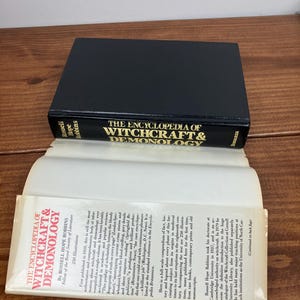 Może przedstawiać: Twarda oprawa książki zatytułowana "The Encyclopedia of Witchcraft & Demonology" ze złotymi literami na czarnym tle. Książka jest otwarta, ukazując tekst. Jest to pozycja referencyjna o czarownictwie.