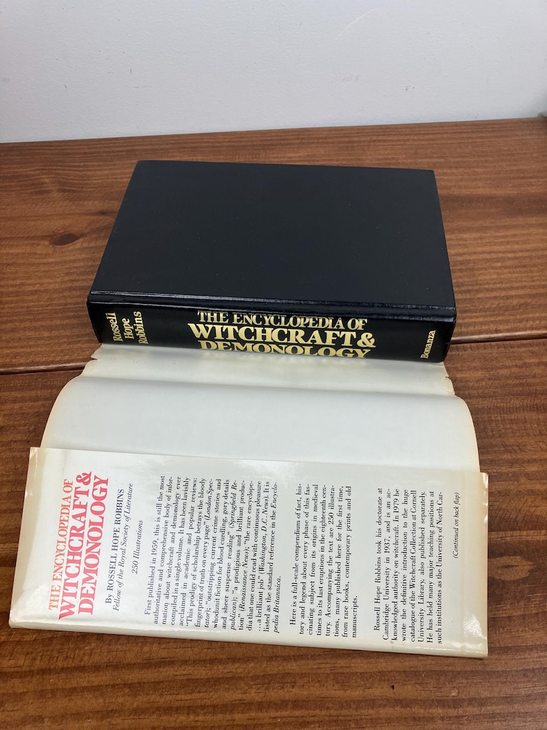 Może przedstawiać: Twarda oprawa książki zatytułowana "The Encyclopedia of Witchcraft & Demonology" ze złotymi literami na czarnym tle. Książka jest otwarta, ukazując tekst. Jest to pozycja referencyjna o czarownictwie.