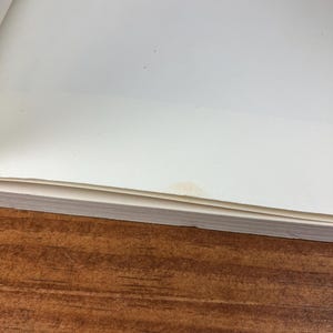 May include: A stack of white paper pages with a small stain on the top right corner, resting on a brown wooden surface. The paper is slightly angled, revealing the edges of the pages.