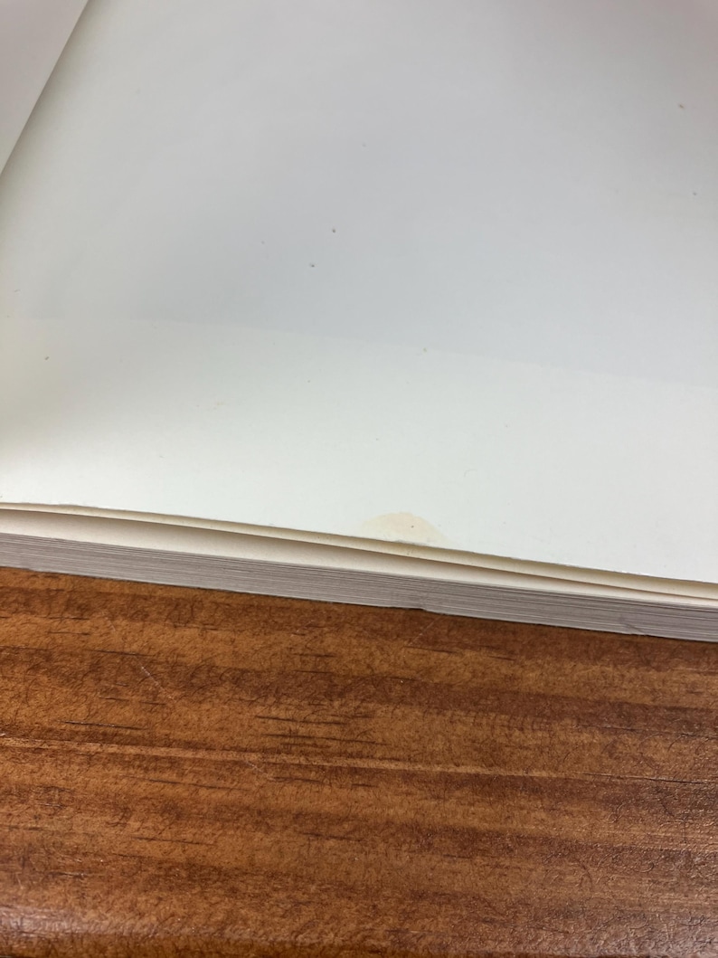 May include: A stack of white paper pages with a small stain on the top right corner, resting on a brown wooden surface. The paper is slightly angled, revealing the edges of the pages.