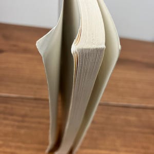 May include: A hardback book with off-white pages, partially open. The spine is visible, and the pages are slightly yellowed. The book is on a wooden surface.