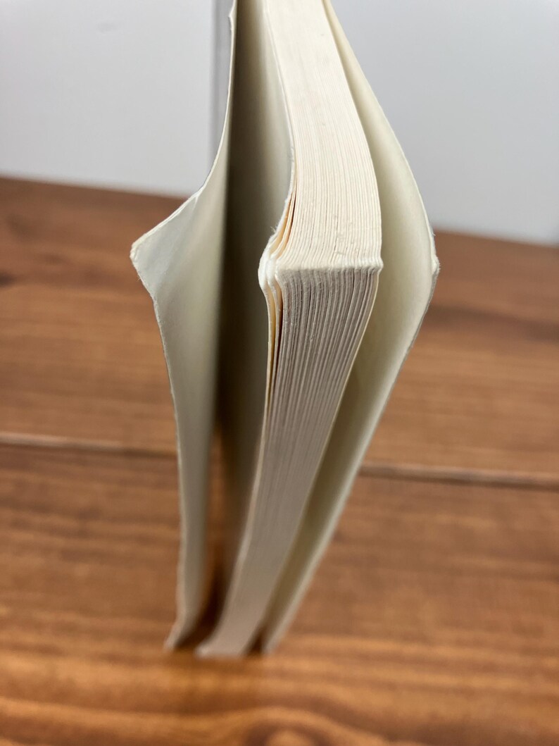 May include: A hardback book with off-white pages, partially open. The spine is visible, and the pages are slightly yellowed. The book is on a wooden surface.