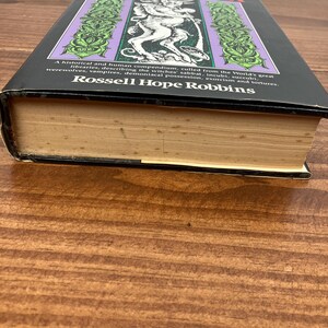 Może przedstawiać: Twarda oprawa książki zatytułowanej "The Encyclopedia of Witchcraft & Demonology" autorstwa Rossella Hope Robbinsa. Okładka przedstawia fioletowo-zielony wz&oacute;r z ilustracją demona. Książka spoczywa na drewnianej powierzchni.