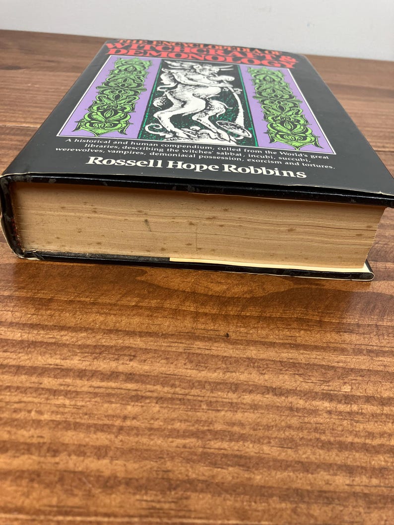 Może przedstawiać: Twarda oprawa książki zatytułowanej "The Encyclopedia of Witchcraft & Demonology" autorstwa Rossella Hope Robbinsa. Okładka przedstawia fioletowo-zielony wz&oacute;r z ilustracją demona. Książka spoczywa na drewnianej powierzchni.