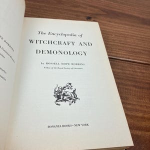 Może przedstawiać: Otwarta książka zatytułowana "The Encyclopedia of Witchcraft and Demonology" autorstwa Rossella Hope Robbinsa. Tytuł książki jest pogrubioną czcionką szeryfową. Logo Bonanza Books znajduje się na dole. Książka jest otwarta na drewnianej powierzchni.