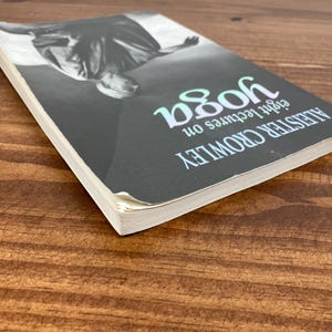 May include: A book titled "Eight Lectures on Yoga" by Aleister Crowley. The cover is black with a grayscale image and the word "yoga" in teal. The book is on a wooden surface.