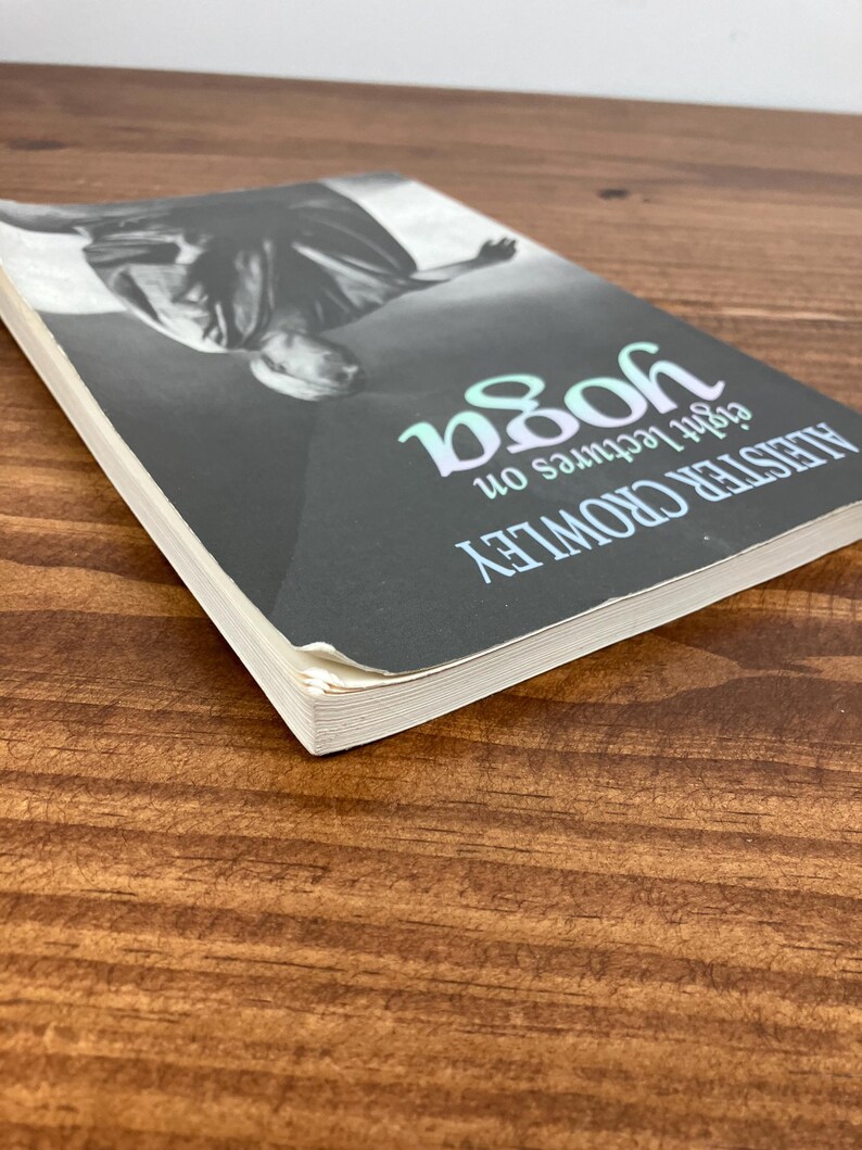 May include: A book titled "Eight Lectures on Yoga" by Aleister Crowley. The cover is black with a grayscale image and the word "yoga" in teal. The book is on a wooden surface.