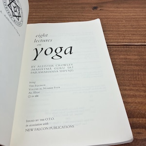 May include: An open book titled "eight lectures on yoga" by Aleister Crowley. The book has white pages with black text and a circular emblem on the top left. The book is open on a wooden surface.