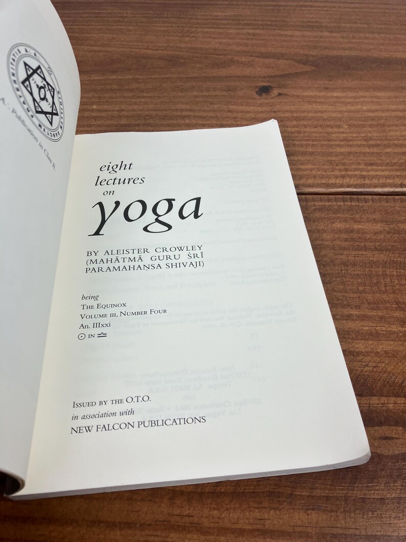 May include: An open book titled "eight lectures on yoga" by Aleister Crowley. The book has white pages with black text and a circular emblem on the top left. The book is open on a wooden surface.