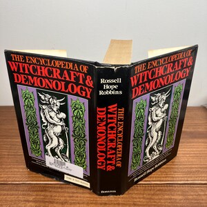 Może przedstawiać: Otwarta książka zatytułowana "THE ENCYCLOPEDIA OF WITCHCRAFT & DEMONOLOGY". Okładka jest czarna z czerwonym i białym tekstem. Zawiera ilustracje postaci i dekoracyjne zielone i fioletowe obramowania. Widoczne jest r&oacute;wnież nazwisko autora, Rossell Hope Robbins.