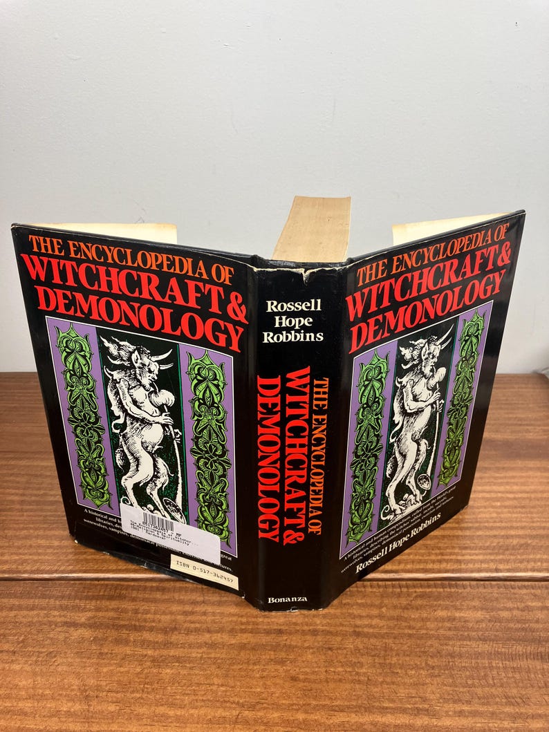 Może przedstawiać: Otwarta książka zatytułowana "THE ENCYCLOPEDIA OF WITCHCRAFT & DEMONOLOGY". Okładka jest czarna z czerwonym i białym tekstem. Zawiera ilustracje postaci i dekoracyjne zielone i fioletowe obramowania. Widoczne jest r&oacute;wnież nazwisko autora, Rossell Hope Robbins.