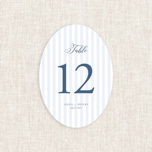 May include: An oval table number card with a blue and white striped background. The word "Table" is written in a script font above the number "12" in a bold, sans-serif font. The names "ANNA + HENRY" and the date "July 27, 2027" are printed at the bottom.