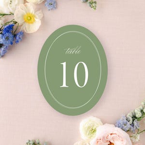May include: An oval, sage green table number card with the number 10 in white script. The card is surrounded by a white oval border. The background is a light pink with floral arrangements and candles.