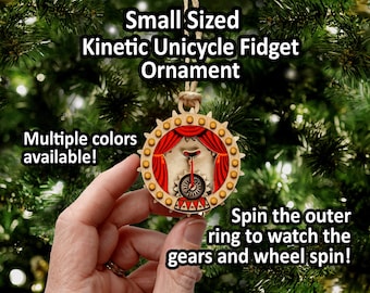May include: A circular wooden kinetic unicycle fidget ornament featuring a red curtain backdrop, a unicycle graphic, and a gear-shaped outer ring. The text reads: Small Sized Kinetic Unicycle Fidget Ornament, Multiple colors available!, Spin the outer ring to watch the gears and wheel spin!