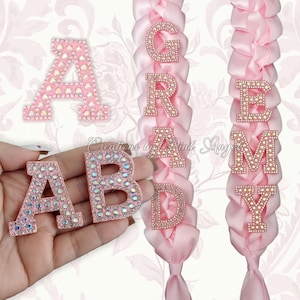 May include: A hand holds two pink letter patches, A and B, covered in iridescent rhinestones. Beside them are two long, braided pink satin ribbons, each adorned with vertical rows of rhinestone-encrusted letters spelling GRAD and EMY.