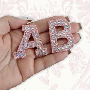 May include: Two light pink fabric letter patches, A and B, decorated with iridescent rhinestones. Each letter features a border of small clear crystals and a center filled with larger, shimmering, multi-colored faceted stones.