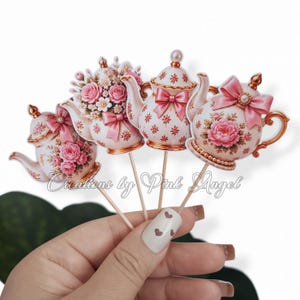 May include: Four decorative teapot-shaped cupcake toppers featuring white porcelain designs with pink floral patterns, gold trim, and pink bows. Each topper is attached to a thin wooden stick. The text Creations by Pink Angel is visible.