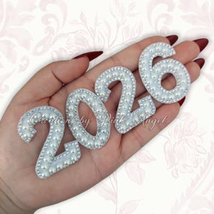 May include: A set of four individual number patches forming the year 2026. Each number is crafted with a white base, bordered by small clear rhinestones, and filled with a single row of round white faux pearls.