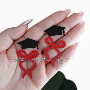 May include: Two black graduation caps with red bows. The image also includes the text "Creations by Pink Angel" in cursive font. The items are held in a person's hand.