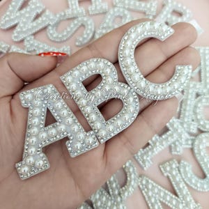 May include: Three white letter patches A, B, and C, each embellished with a border of small clear rhinestones and a center row of white faux pearls. The letters are arranged together, and the text Creations by Pink Angel is visible.