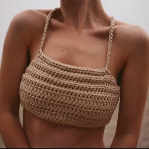 May include: A beige crochet crop top with braided straps. The top features a horizontal ribbed pattern and a square neckline. The neutral color and textured design create a bohemian aesthetic, suitable for beachwear or summer outfits.