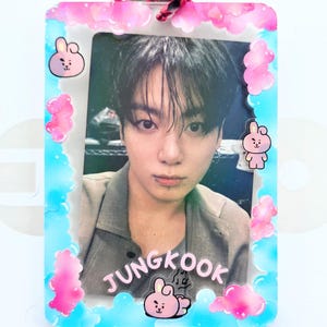 May include: A rectangular acrylic photo card holder featuring a blue border with pink cloud accents and small pink cartoon rabbit characters. Inside is a portrait of a person with dark hair and a lip piercing, with the name JUNGKOOK printed below.