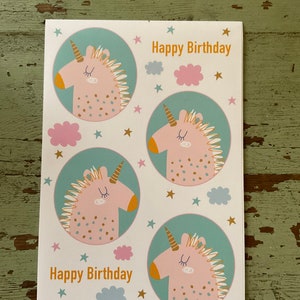 Puede incluir: Una tarjeta blanca con un dise&ntilde;o de unicornio turquesa y rosa. La tarjeta tiene el texto "Happy Birthday" escrito dos veces. La tarjeta est&aacute; decorada con estrellas doradas y nubes rosas y azules.