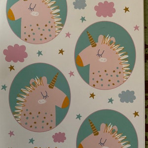 Puede incluir: Una tarjeta blanca con un esquema de color turquesa, verde y rosa. La tarjeta presenta cuatro unicornios rosas con cuernos y crines dorados. Los unicornios est&aacute;n rodeados de estrellas doradas y nubes rosas y azules. El texto "Happy Birthday" est&aacute; escrito en letras doradas en la parte superior e inferior de la tarjeta.