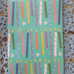 May include: A teal blue wrapping paper with a repeating pattern of colorful birthday candles with white polka dots, stripes, and other patterns. The candles have a yellow flame on top. There are also small white flowers scattered throughout the pattern.