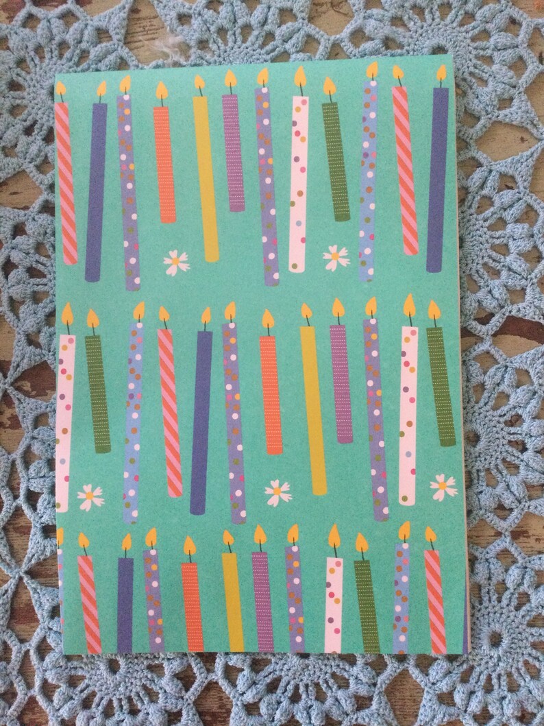 May include: A teal blue wrapping paper with a repeating pattern of colorful birthday candles with white polka dots, stripes, and other patterns. The candles have a yellow flame on top. There are also small white flowers scattered throughout the pattern.