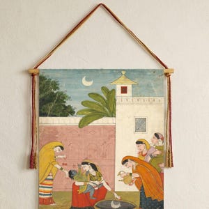May include: An Indian miniature painting depicting a scene with figures in traditional attire. The artwork features a woman holding a baby, surrounded by other women, with cows in the foreground. The painting is mounted on wooden dowels and hung with a cord.